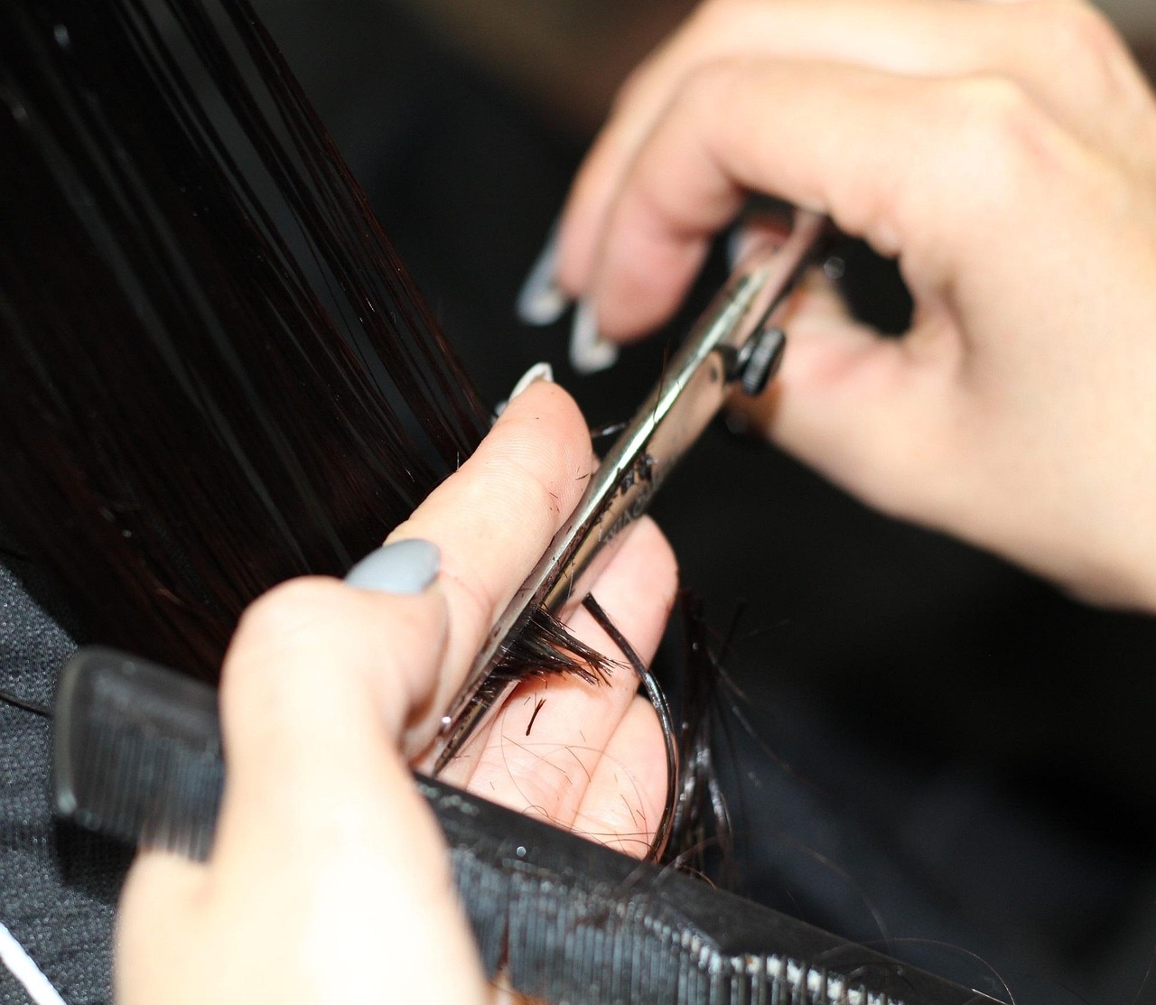 Close-up of a stylist's hands with painted nails trimming a client's dark hair with scissors and a comb