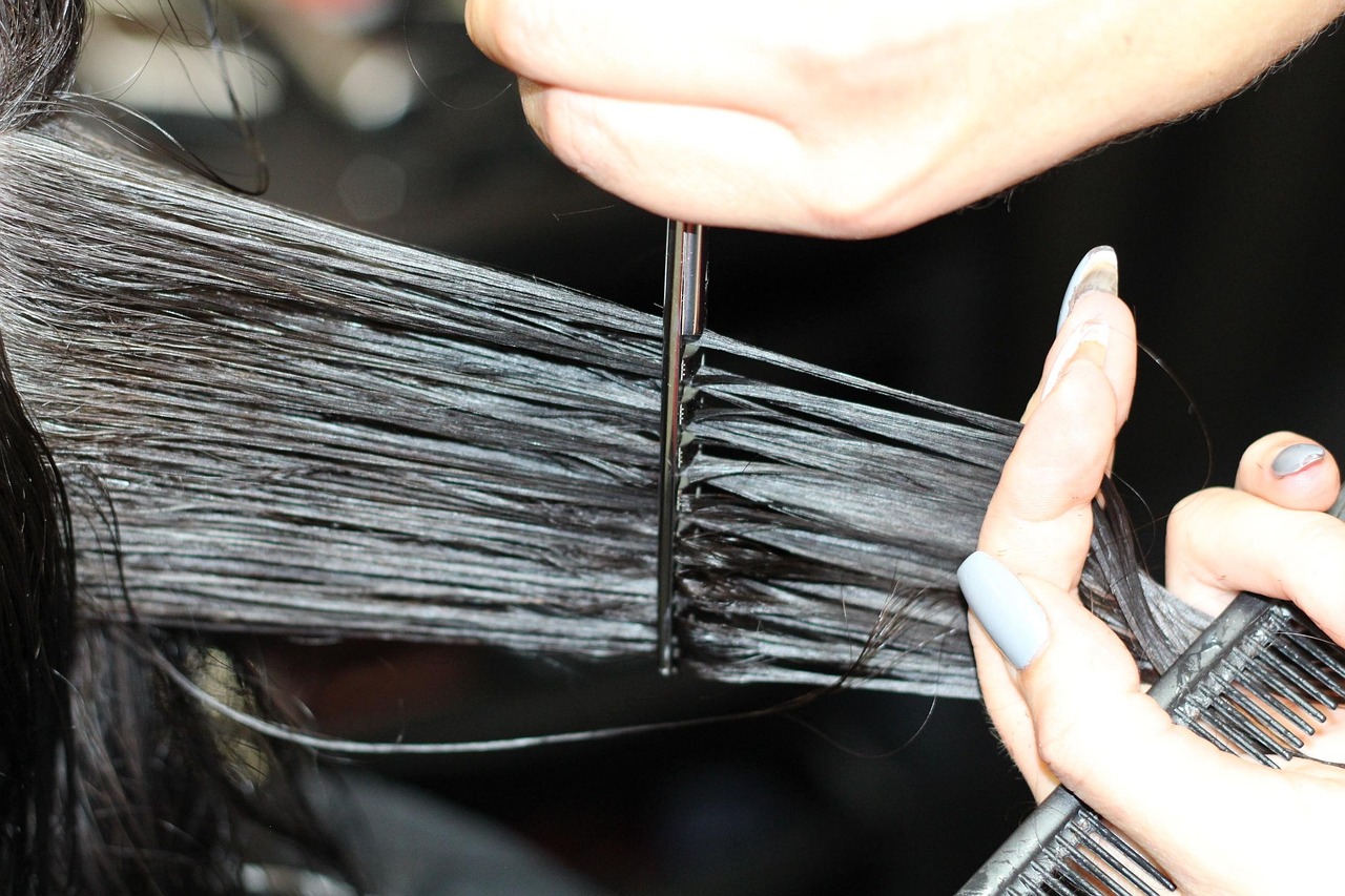Close-up of a stylist's hands holding a comb and trimming a section of a client's long dark hair with scissors